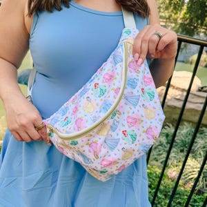 May include: A white fanny pack featuring a repeating pattern of various cartoon princesses in colorful dresses. The bag has a gold-toned zipper closure and an adjustable light-colored strap. The design includes multiple figures in pink, blue, and yellow.