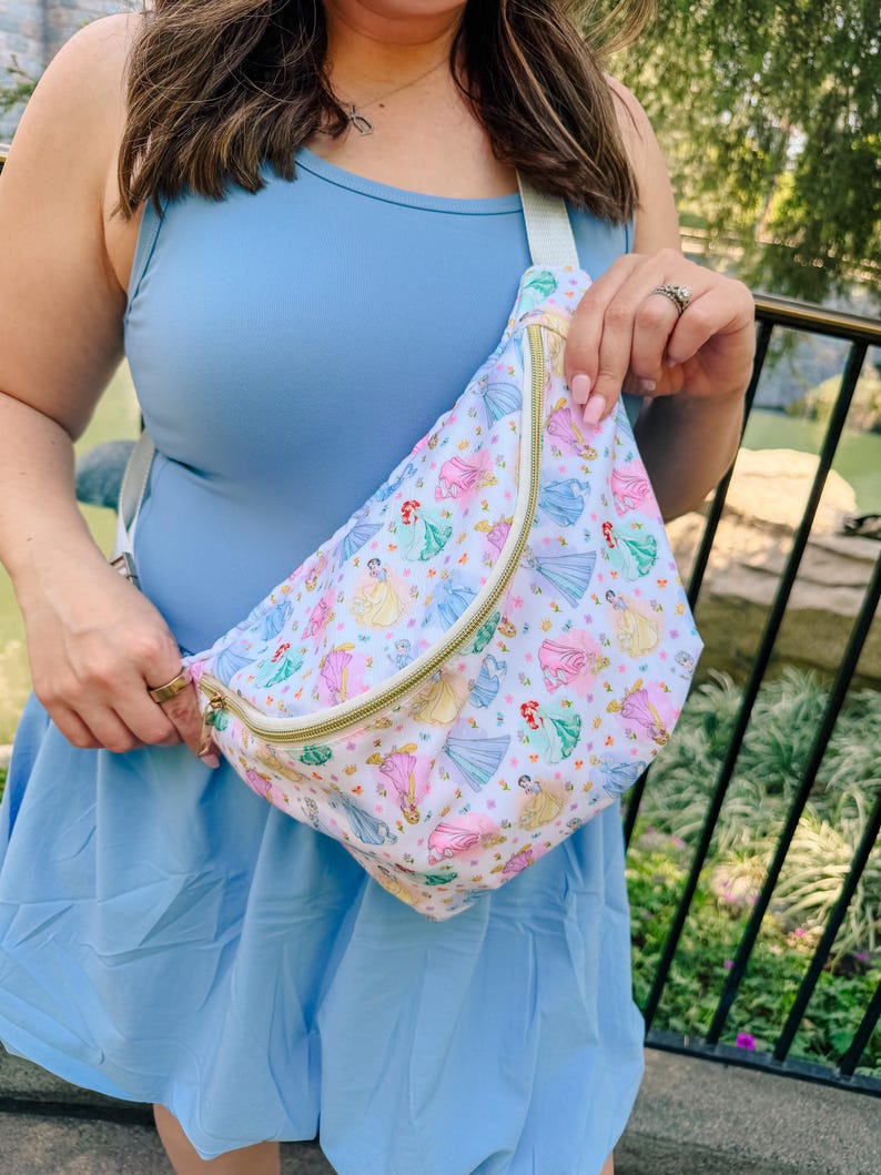May include: A white fanny pack featuring a repeating pattern of various cartoon princesses in colorful dresses. The bag has a gold-toned zipper closure and an adjustable light-colored strap. The design includes multiple figures in pink, blue, and yellow.
