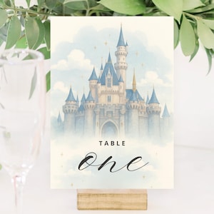 Puede incluir: Una tarjeta con el número de mesa con una ilustración en acuarela de un castillo de cuento de hadas con torretas azules y un fondo de cielo azul claro. La tarjeta dice "TABLE" encima de la palabra "one" en una elegante escritura. La tarjeta está sujeta por un pequeño bloque de madera.