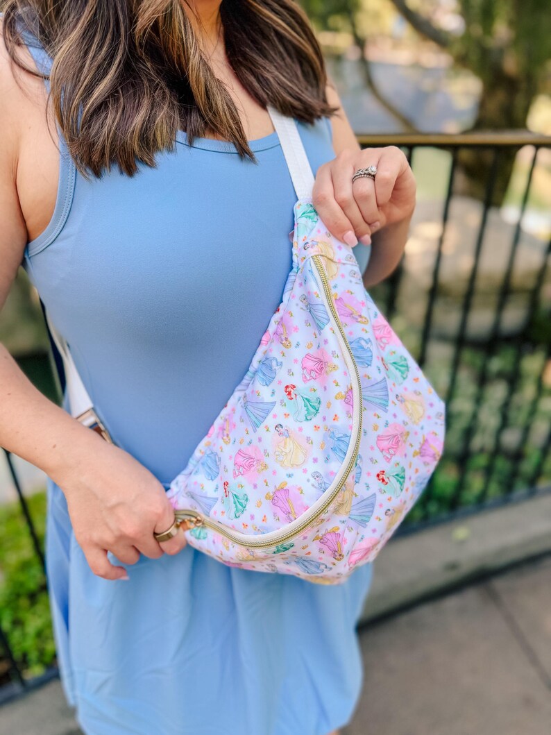 May include: A white crossbody sling bag featuring a repeating pattern of various colorful cartoon princesses in ballgowns. The bag has a gold-toned zipper closure and a wide white adjustable strap, worn across the chest over a light blue sleeveless dress.