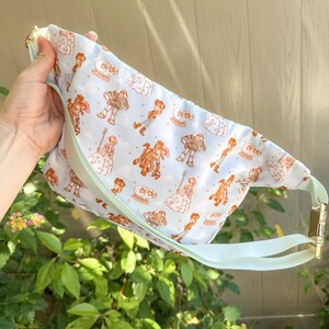 May include: A light blue fanny pack with a repeating pattern of cartoon characters in shades of brown and white. The bag has a light green strap and gold-colored hardware. The bag is held up by a hand.