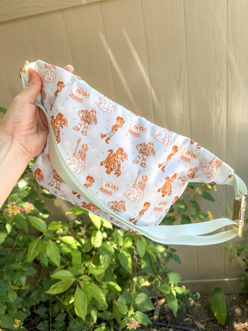 May include: A light blue fanny pack with a repeating pattern of cartoon characters in shades of brown and white. The bag has a light green strap and gold-colored hardware. The bag is held up by a hand.