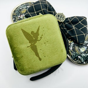 May include: A green velvet jewelry case with a silhouette of Tinkerbell. The case has a black wrist strap and a zipper closure. Two other small cases are visible, one with a floral pattern and the other with a black quilted design.