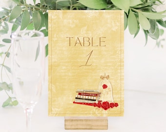 May include: A table number card with a yellow background and the words "TABLE 1" in brown. The card features a stack of books, a glass bell with a rose, and red roses at the bottom. The card is held by a wooden stand.