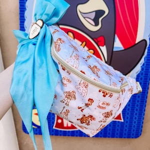 May include: A light blue fanny pack with a zipper closure and a pattern of cartoon characters. The fanny pack is tied with a light blue ribbon and a wooden tag that says "ANDY".
