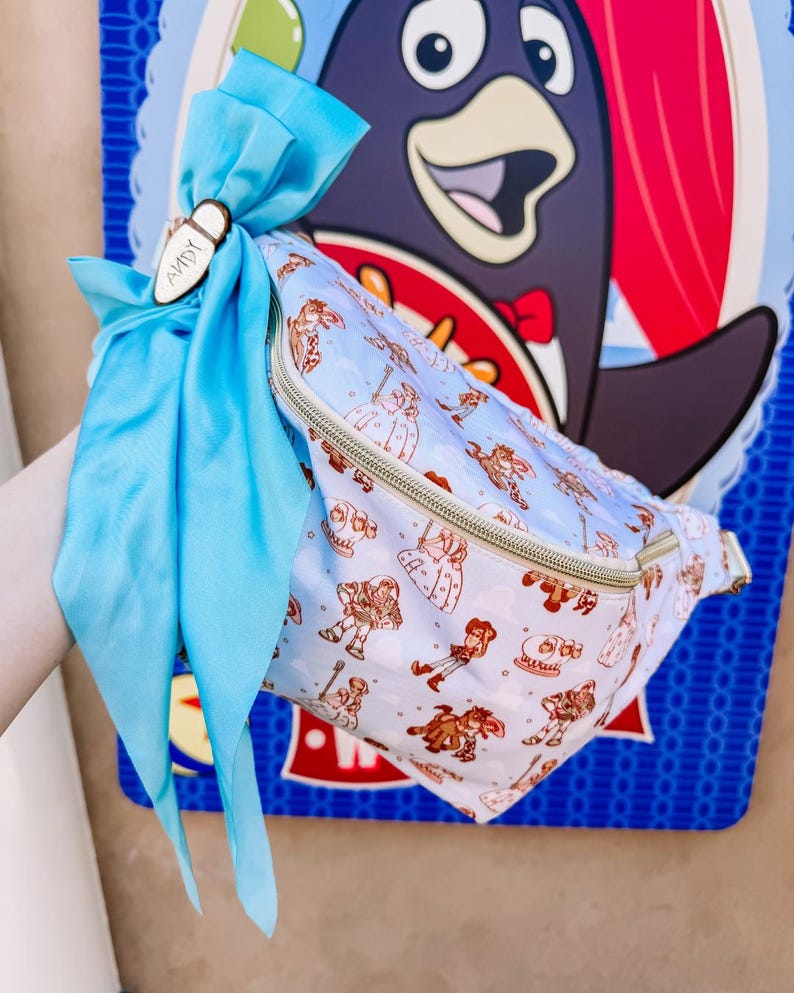 May include: A light blue fanny pack with a zipper closure and a pattern of cartoon characters. The fanny pack is tied with a light blue ribbon and a wooden tag that says "ANDY".