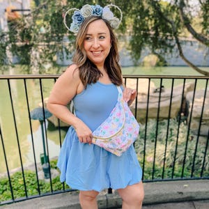 May include: A light blue sleeveless dress with a flared skirt, paired with a white fanny pack featuring a colorful pattern of cartoon characters and a gold zipper, and a headband adorned with light blue and white fabric roses.