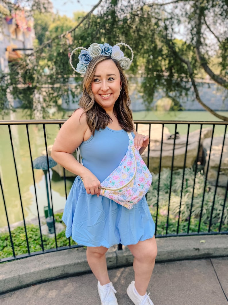 May include: A light blue sleeveless dress with a flared skirt, paired with a white fanny pack featuring a colorful pattern of cartoon characters and a gold zipper, and a headband adorned with light blue and white fabric roses.