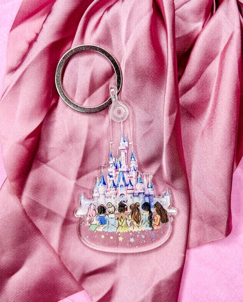 Puede incluir: Un llavero de acr&iacute;lico transparente con una ilustraci&oacute;n colorida del castillo de Cenicienta con siete princesas Disney de pie frente a &eacute;l. Las princesas llevan sus vestidos caracter&iacute;sticos y tienen detalles brillantes.