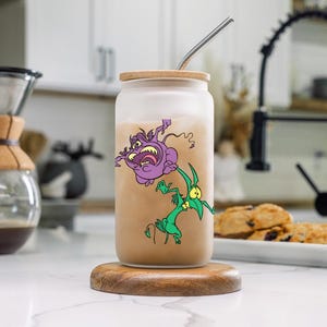 Peut inclure: Un gobelet en verre givré avec un couvercle brun et une paille en acier inoxydable. Le gobelet est illustré de deux créatures de dessin animé, l'une violette et l'autre verte. Le gobelet est posé sur un dessous de verre en bois.