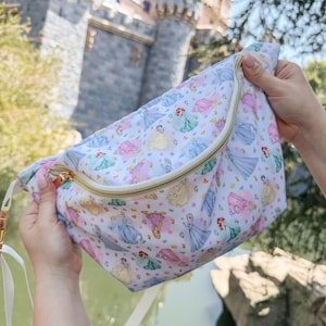 May include: A white fanny pack featuring an all-over print of various cartoon princesses in colorful dresses, accented with small floral motifs. The bag includes a gold-toned zipper closure and a white adjustable strap for secure carrying.