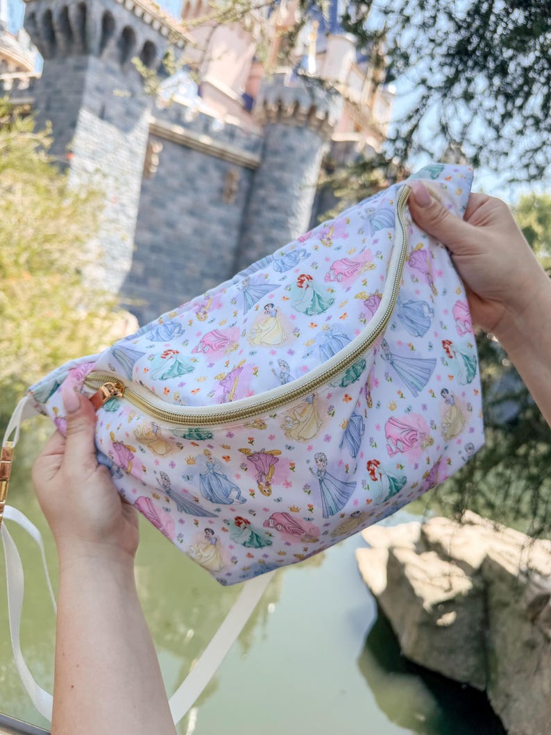 May include: A white fanny pack featuring an all-over print of various cartoon princesses in colorful dresses, accented with small floral motifs. The bag includes a gold-toned zipper closure and a white adjustable strap for secure carrying.