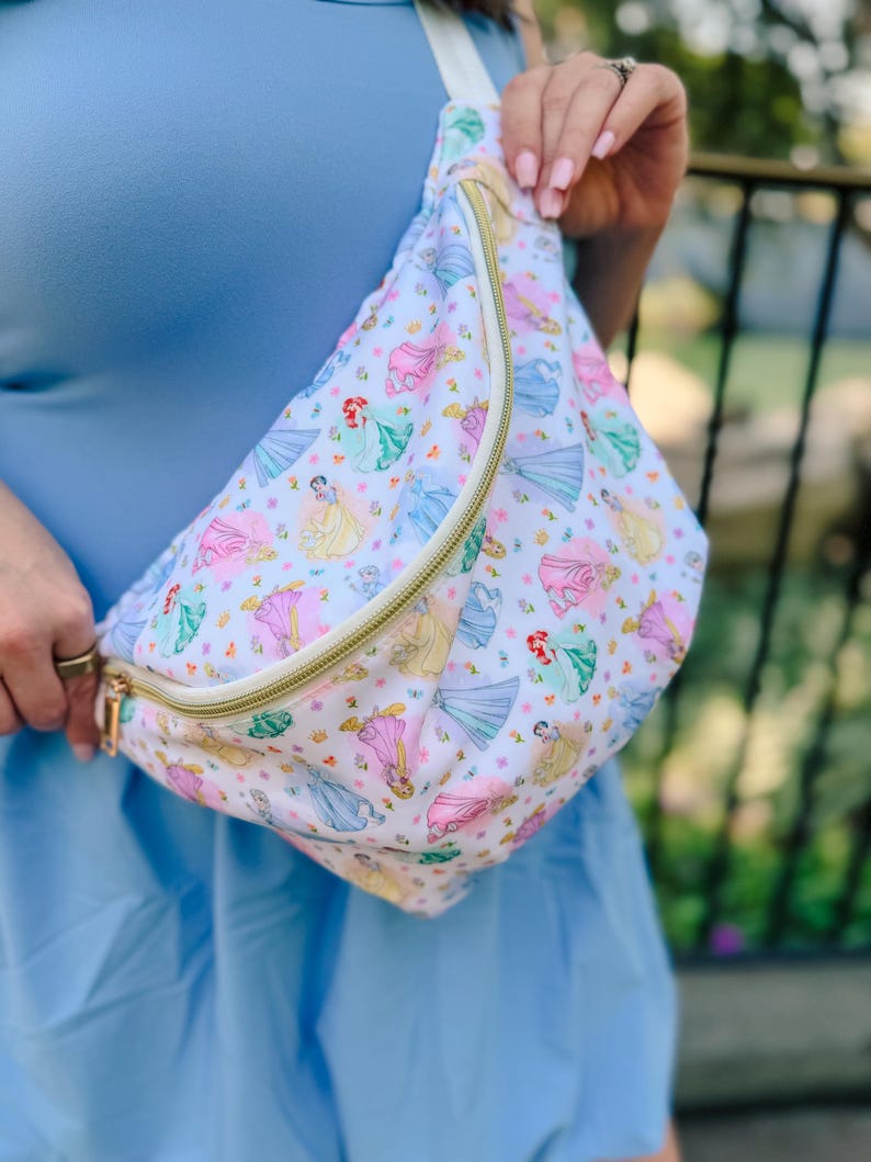 May include: A white fanny pack featuring a repeating pattern of various cartoon princesses in colorful dresses, accented with small yellow flowers. The bag includes a gold-toned zipper closure and an adjustable white strap for secure carrying.