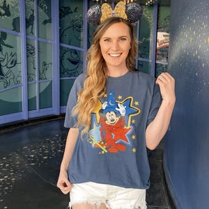 May include: A woman wearing a blue t-shirt with a Mickey Mouse graphic and white denim shorts. She is wearing a pair of Minnie Mouse ears with gold glitter.