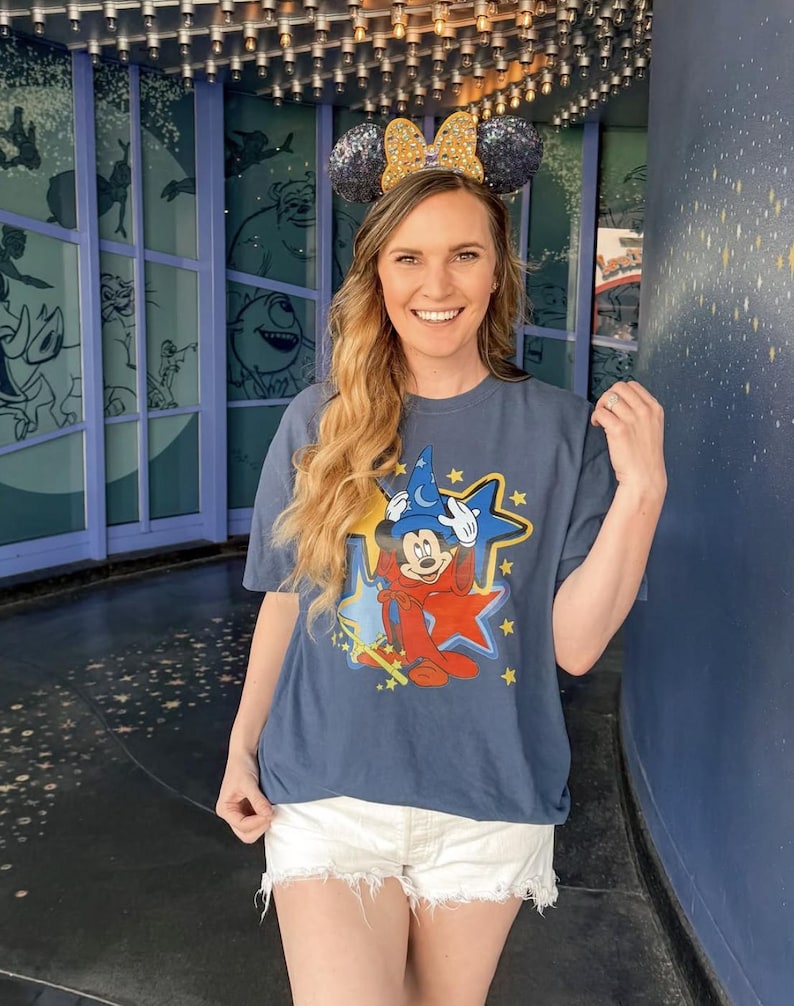 May include: A woman wearing a blue t-shirt with a Mickey Mouse graphic and white denim shorts. She is wearing a pair of Minnie Mouse ears with gold glitter.