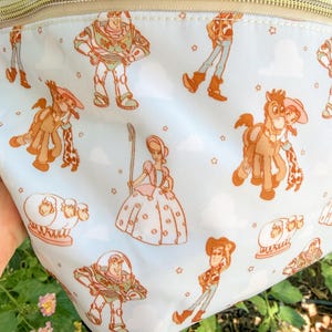May include: A white fanny pack with a gold zipper and a repeating pattern of Toy Story characters in orange and brown. The characters include Buzz Lightyear, Woody, Jessie, and Bo Peep, with small stars scattered throughout.