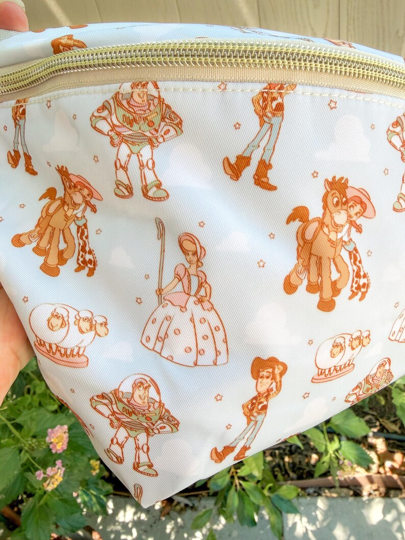 May include: A white fanny pack with a gold zipper and a repeating pattern of Toy Story characters in orange and brown. The characters include Buzz Lightyear, Woody, Jessie, and Bo Peep, with small stars scattered throughout.