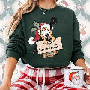May include: A dark green sweatshirt featuring a cartoon dog wearing a Santa hat and holding a sign that reads "to: santa." The sweatshirt is paired with red and white plaid pajama pants. A white mug with a Santa Claus design is also visible.