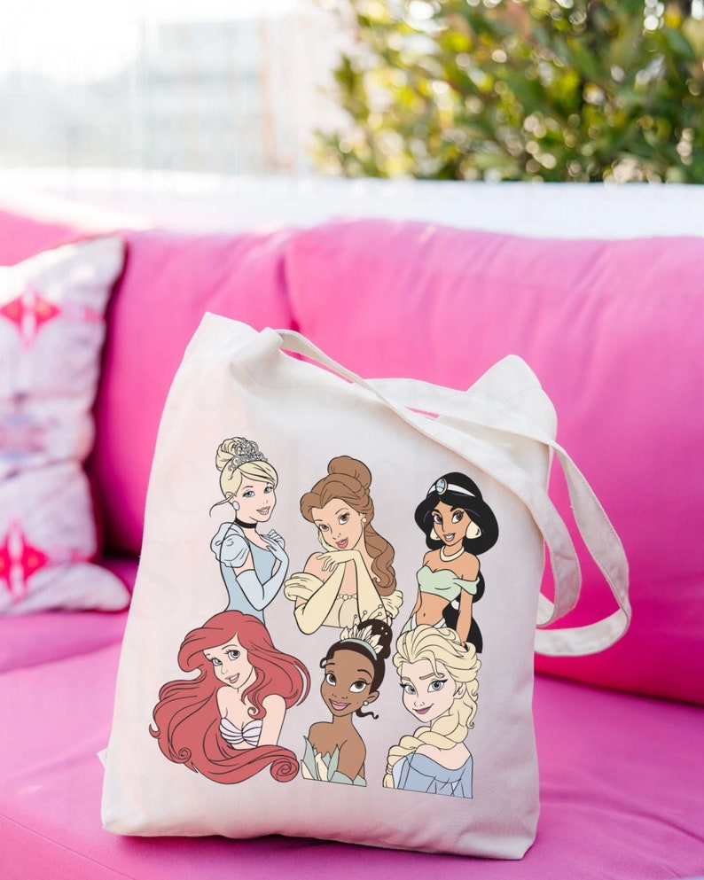May include: A white canvas tote bag with a colorful print of Disney princesses. The princesses include Cinderella, Belle, Jasmine, Ariel, Tiana, and Elsa.