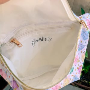 May include: A white interior bag with a gold zipper and a zippered pocket featuring the black text Ever After Design. The exterior fabric displays a repeating pattern of colorful princess figures in various dresses against a white background.