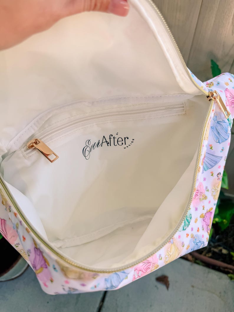 May include: A white interior bag with a gold zipper and a zippered pocket featuring the black text Ever After Design. The exterior fabric displays a repeating pattern of colorful princess figures in various dresses against a white background.