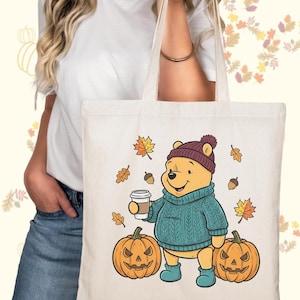 May include: Cream-colored tote bag featuring a cartoon bear in a teal sweater, burgundy hat, and blue boots, holding a coffee cup. Two carved pumpkins and autumn leaves surround the bear. A fall-themed design perfect for carrying groceries or books.