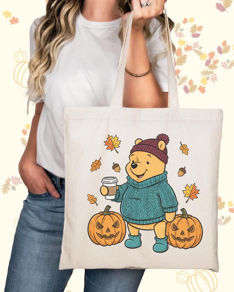 May include: Cream-colored tote bag featuring a cartoon bear in a teal sweater, burgundy hat, and blue boots, holding a coffee cup. Two carved pumpkins and autumn leaves surround the bear. A fall-themed design perfect for carrying groceries or books.