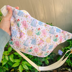 May include: A white fabric fanny pack featuring a repeating pattern of various cartoon princesses in colorful gowns, including blue, pink, yellow, and green dresses, accented with small gold crowns and tiny pink flowers, complete with an adjustable beige strap.