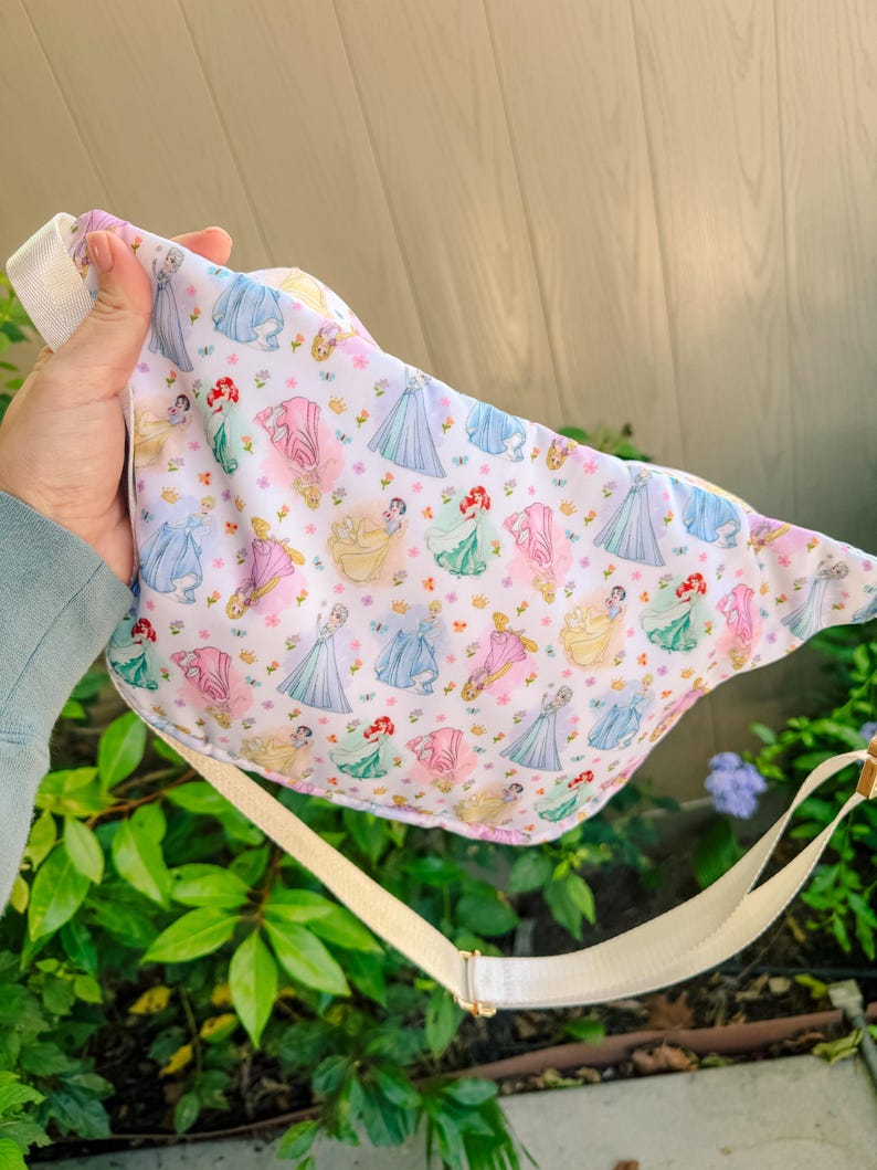 May include: A white fabric fanny pack featuring a repeating pattern of various cartoon princesses in colorful gowns, including blue, pink, yellow, and green dresses, accented with small gold crowns and tiny pink flowers, complete with an adjustable beige strap.