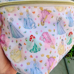 May include: A white fanny pack featuring a repeating pattern of various animated princesses in colorful gowns, small gold crowns, and tiny flowers. The bag has a gold-toned zipper closure along the top edge and a smooth, lightweight fabric construction.