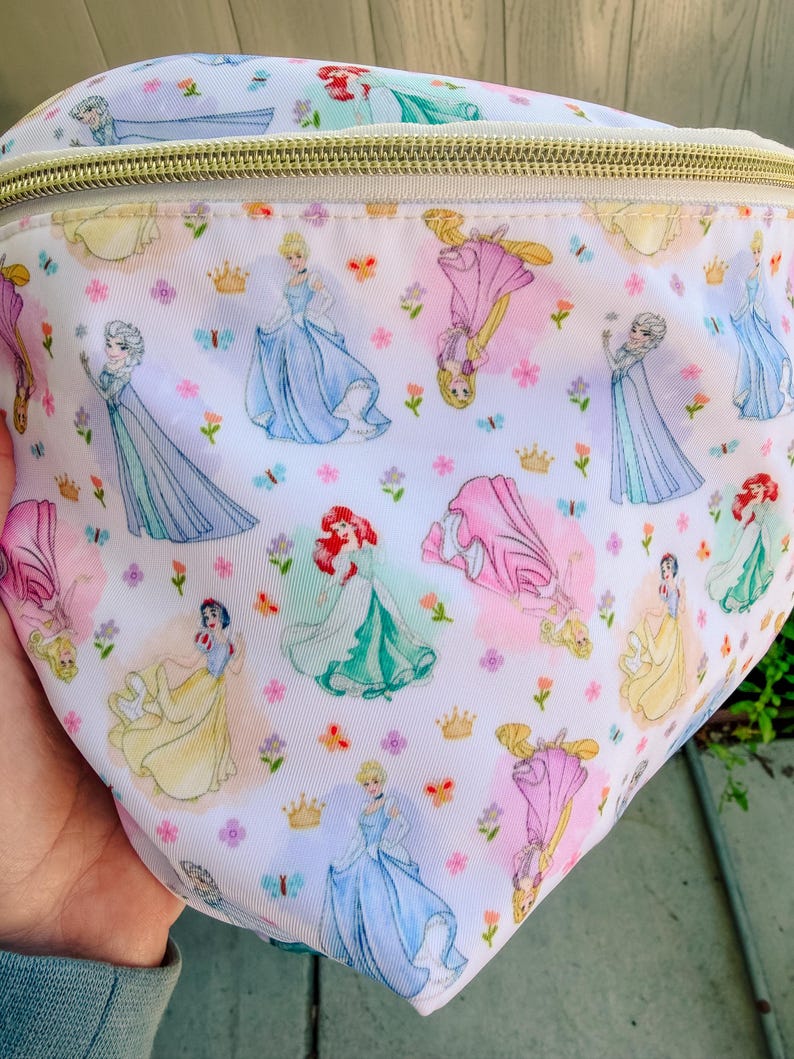 May include: A white fanny pack featuring a repeating pattern of various animated princesses in colorful gowns, small gold crowns, and tiny flowers. The bag has a gold-toned zipper closure along the top edge and a smooth, lightweight fabric construction.