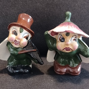 May include: Two ceramic figurines of whimsical characters. One is wearing a brown hat and green clothing, holding a black instrument. The other is wearing a pink and white hat and green clothing. Both have painted faces with large eyes.