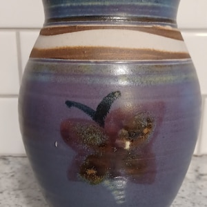 May include: A ceramic vase with a purple, brown, and white glaze. The vase has a wide mouth and a rounded body. It features a stylized flower design in shades of brown and black.
