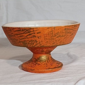 May include: Orange and gold ceramic pedestal bowl with a white interior. The bowl has a textured, wood-grain design.