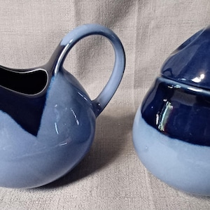 May include: A light blue and navy blue ceramic creamer and sugar bowl set. The creamer has a handle and spout, while the sugar bowl has a lid with a rounded top. The set has a glossy finish.