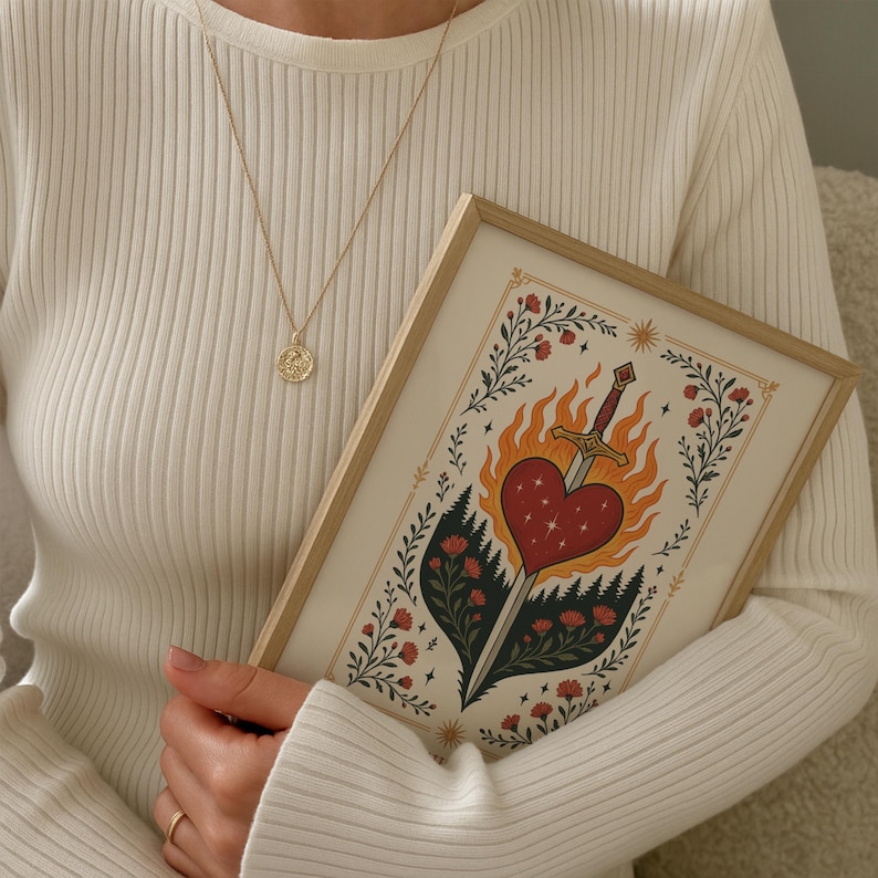Op de afbeelding: Een ingelijste kunstdruk met een hart doorboord door een zwaard, omringd door vlammen en bloemmotieven. De persoon draagt een gouden hangerketting en een witte geribbelde trui.