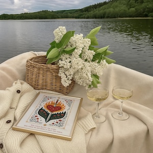 Op de afbeelding: Een picknicksc&egrave;ne met een rieten mand, een ingelijste print en twee champagneglazen. De print toont een hart doorboord door een zwaard, omringd door vlammen en bloemen. Een boeket witte bloemen en een cr&egrave;mekleurig vest completeren de sc&egrave;ne.