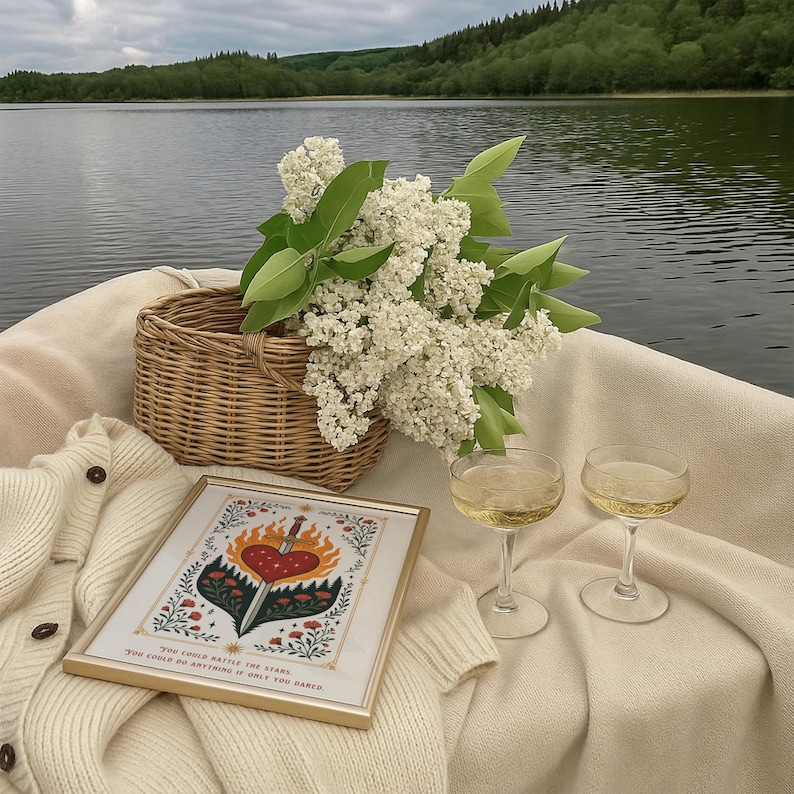 Op de afbeelding: Een picknicksc&egrave;ne met een rieten mand, een ingelijste print en twee champagneglazen. De print toont een hart doorboord door een zwaard, omringd door vlammen en bloemen. Een boeket witte bloemen en een cr&egrave;mekleurig vest completeren de sc&egrave;ne.