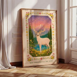 Puede incluir: Una impresión artística enmarcada que presenta un ciervo blanco con grandes astas cerca de un lago y montañas. La obra de arte tiene un borde floral y el texto "You could rattle the stars". La paleta de colores incluye rosa, amarillo, verde y azul.