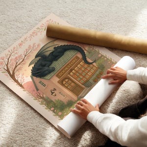 May include: A rolled-up print featuring a whimsical illustration of a bookstore with a dragon perched on the roof. The print is surrounded by cherry blossoms and a light pink border. A second rolled print is visible.