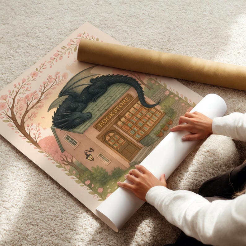 May include: A rolled-up print featuring a whimsical illustration of a bookstore with a dragon perched on the roof. The print is surrounded by cherry blossoms and a light pink border. A second rolled print is visible.