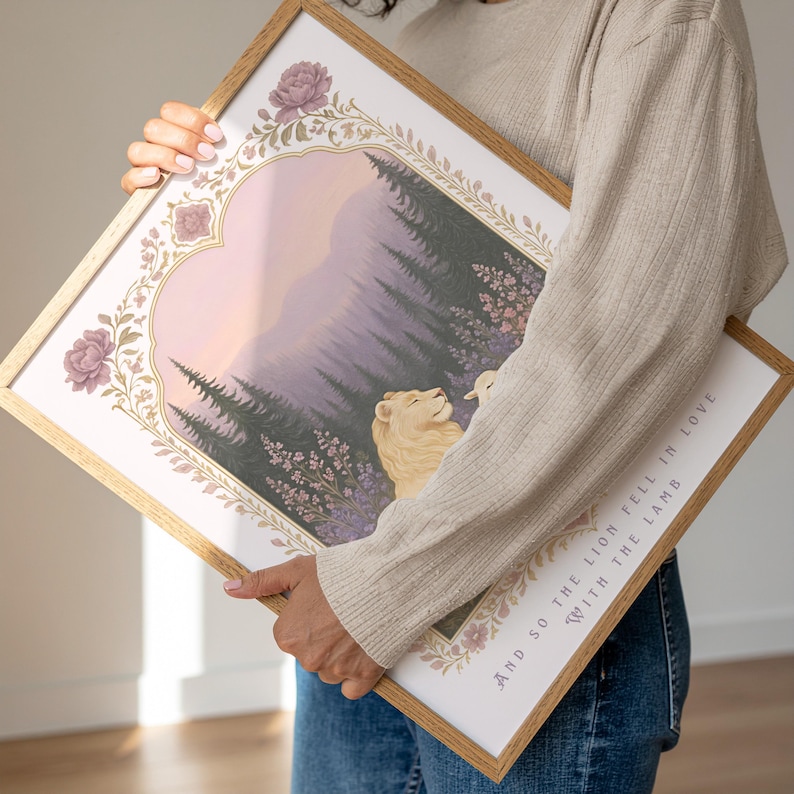 May include: A framed art print featuring a lion and a lamb in a forest scene with a mountain backdrop. The artwork has a soft color palette of lavender, pink, and gold. The frame is a light wood color, and the text "AND SO THE LION FELL IN LOVE WITH THE LAMB" is visible.