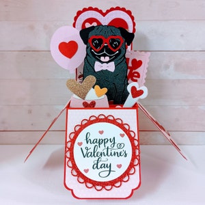 May include: A pop-up card with a black pug wearing heart-shaped sunglasses and a bow tie. The card is decorated with hearts, glitter, and the text "happy Valentine's day".