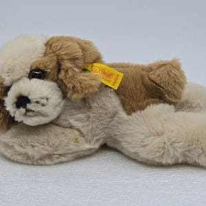 May include: A plush dog toy in shades of tan and brown, lying down. The toy has floppy ears, a black nose, and a yellow tag attached. The dog's fur is soft and fluffy, with a realistic appearance.