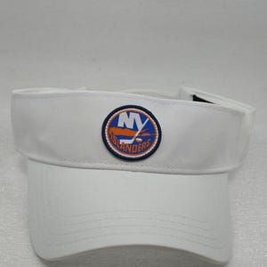 May include: A white visor featuring a circular patch on the front panel. The patch displays the blue and orange New York Islanders logo, which includes the letters NY, a hockey stick, and a puck, all surrounded by a dark blue stitched border.