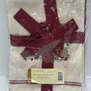 Peut inclure: Un ensemble de serviettes en tissu couleur crème avec un motif de guirlande naturelle imprimé, noué par un ruban rouge foncé. Une étiquette rectangulaire indique Longaberger Nature's Garland Towel Set, numéro 23155, made in China.