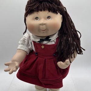 May include: A vintage doll with brown yarn hair and freckles, wearing a red corduroy dress with black buttons and a white blouse. The doll has a small gold heart pendant and is wearing black and white saddle shoes. A plaid bow adorns the top of the head.