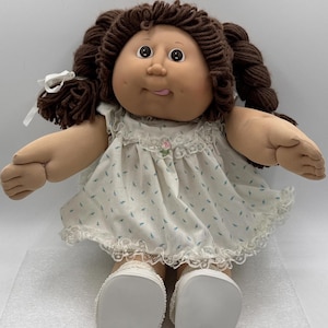 Puede incluir: Muñeca vintage Cabbage Patch Kid con cabello castaño de hilo en coletas. La muñeca viste un vestido blanco con estampado floral azul y ribete de encaje, y zapatos blancos. La muñeca tiene una expresión sonriente.