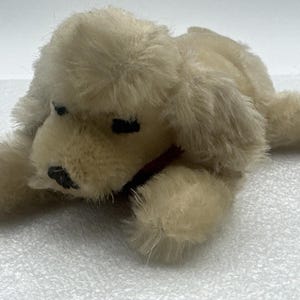 May include: A plush, cream-colored dog toy in a lying down position. The toy has black eyes and a black nose, with a red collar. The fur is soft and fluffy, giving it a cuddly appearance. A classic children's toy.