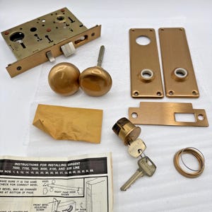 Könnte beinhalten: Eine Sammlung von Vintage-Türbeschlägen aus Messing, darunter ein Einsteckschloss, Türknöpfe, Rosetten und ein Zylinderschloss mit Schlüsseln. Das Set enthält auch Installationsanweisungen. Das Hardware hat ein warmes, metallisches Finish.